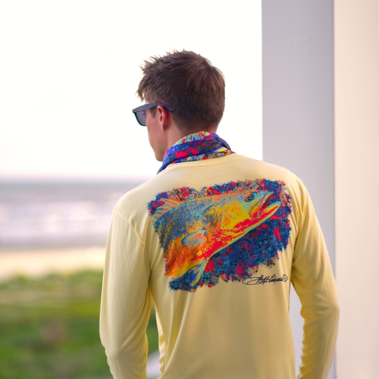 Man wearing Speckled Trout UPF 50+ Fishing Shirt with vivid trout print, standing on a porch with ocean view, highlighting outdoor performance wear.