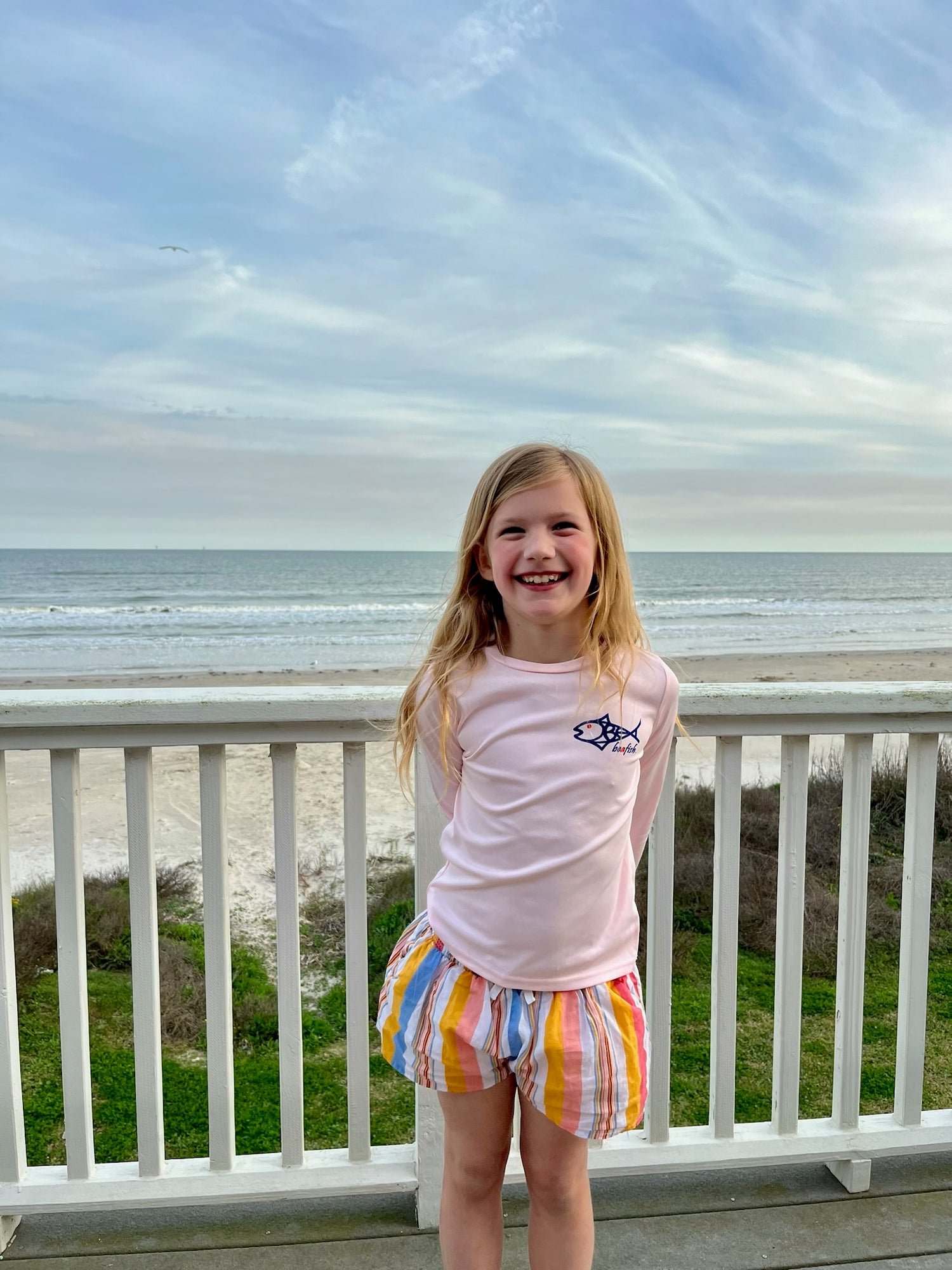 Smiling girl wearing Caribbean Iguana Kids Performance Fishing Shirt UPF 50+ with fish logo, standing on a beachside balcony.