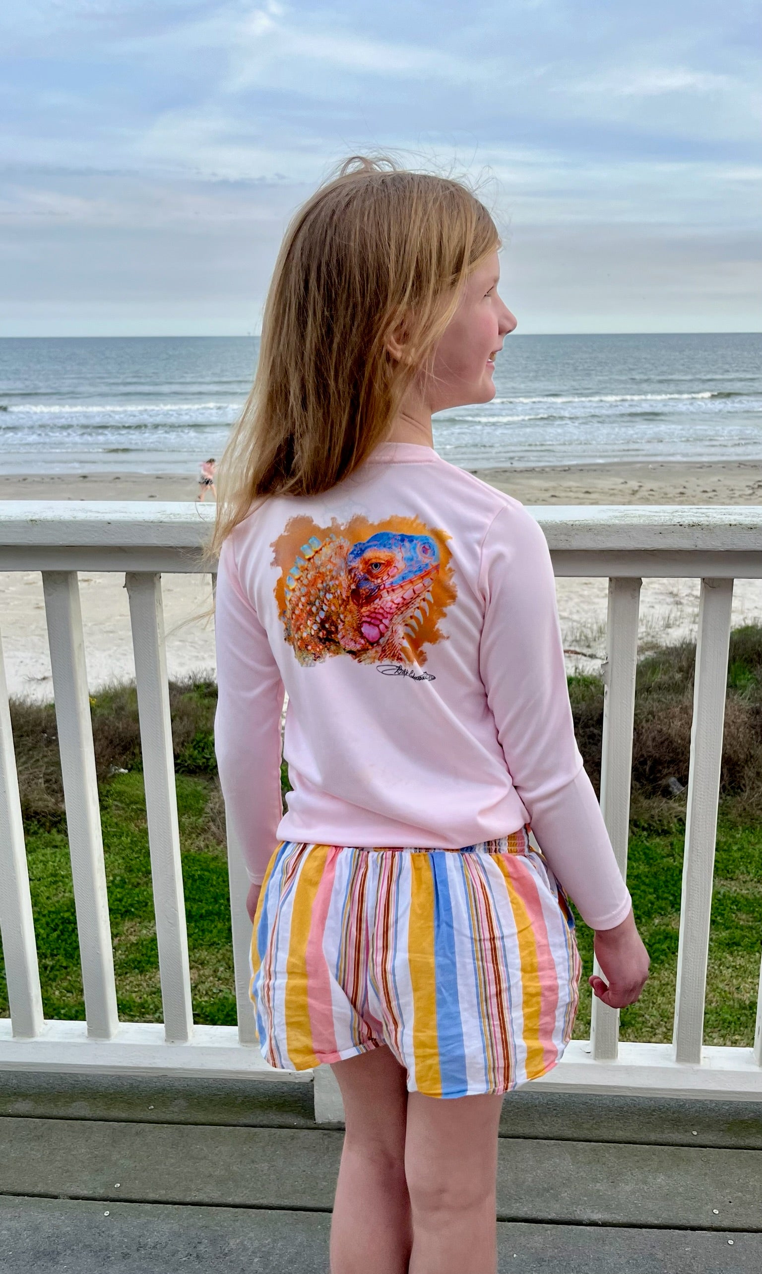 Girl wearing Caribbean Iguana Kids Performance Fishing Shirt UPF 50+ with iguana design on back, standing on beachside deck.