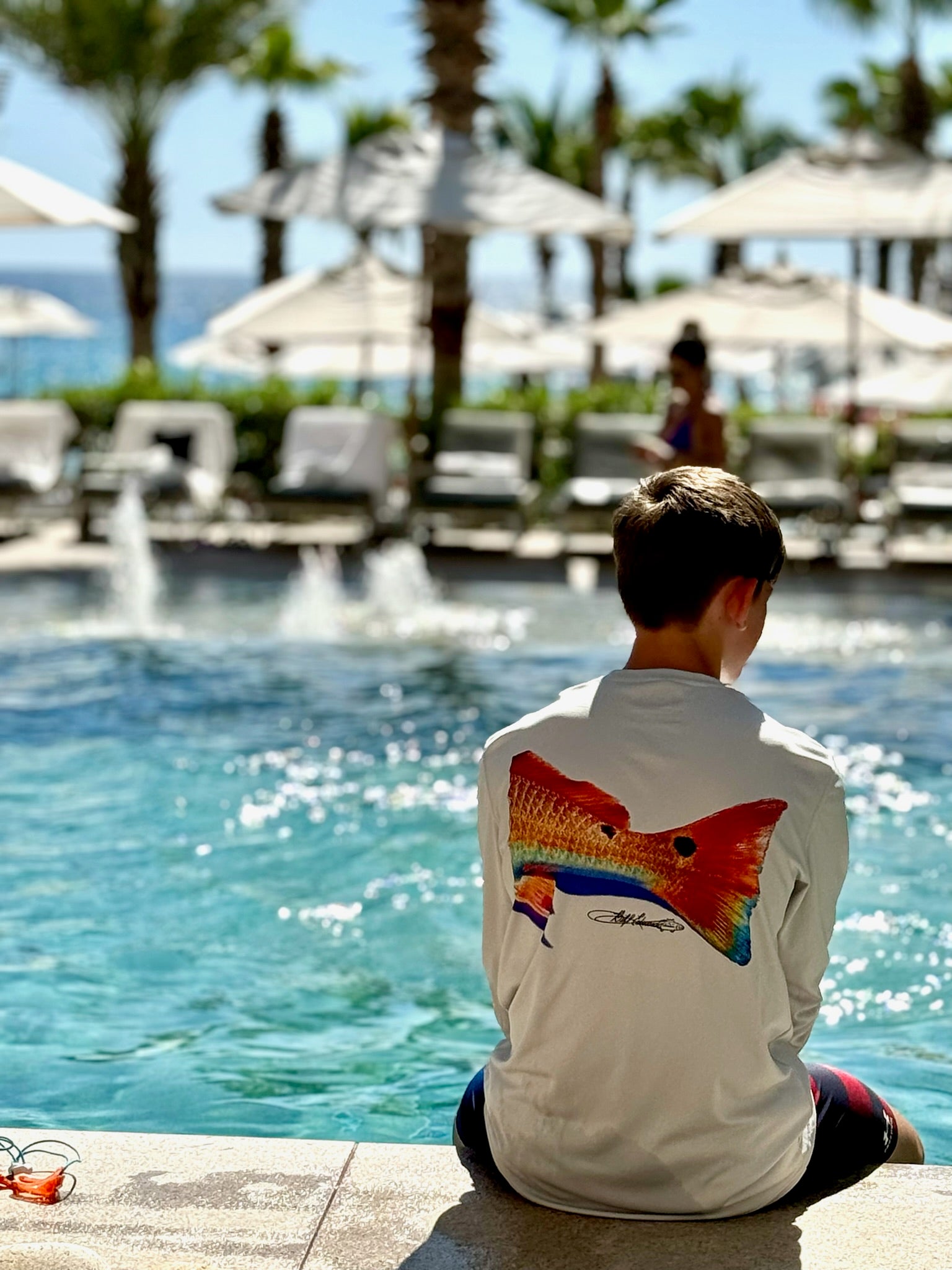 Boy wearing Redfish Design Fishing Shirt with UPF 50 by a resort pool. Long sleeve performance shirt offers sun protection and moisture wicking comfort.