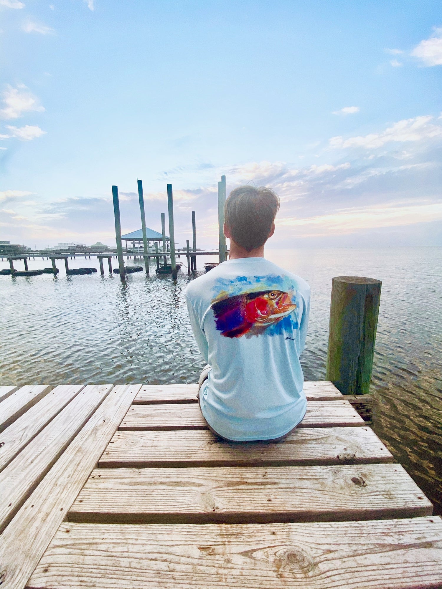 Man sitting on dock at sunset in Rainbow Trout UPF 50+ Shirt, highlighting back trout design and relaxed outdoor lifestyle appeal.