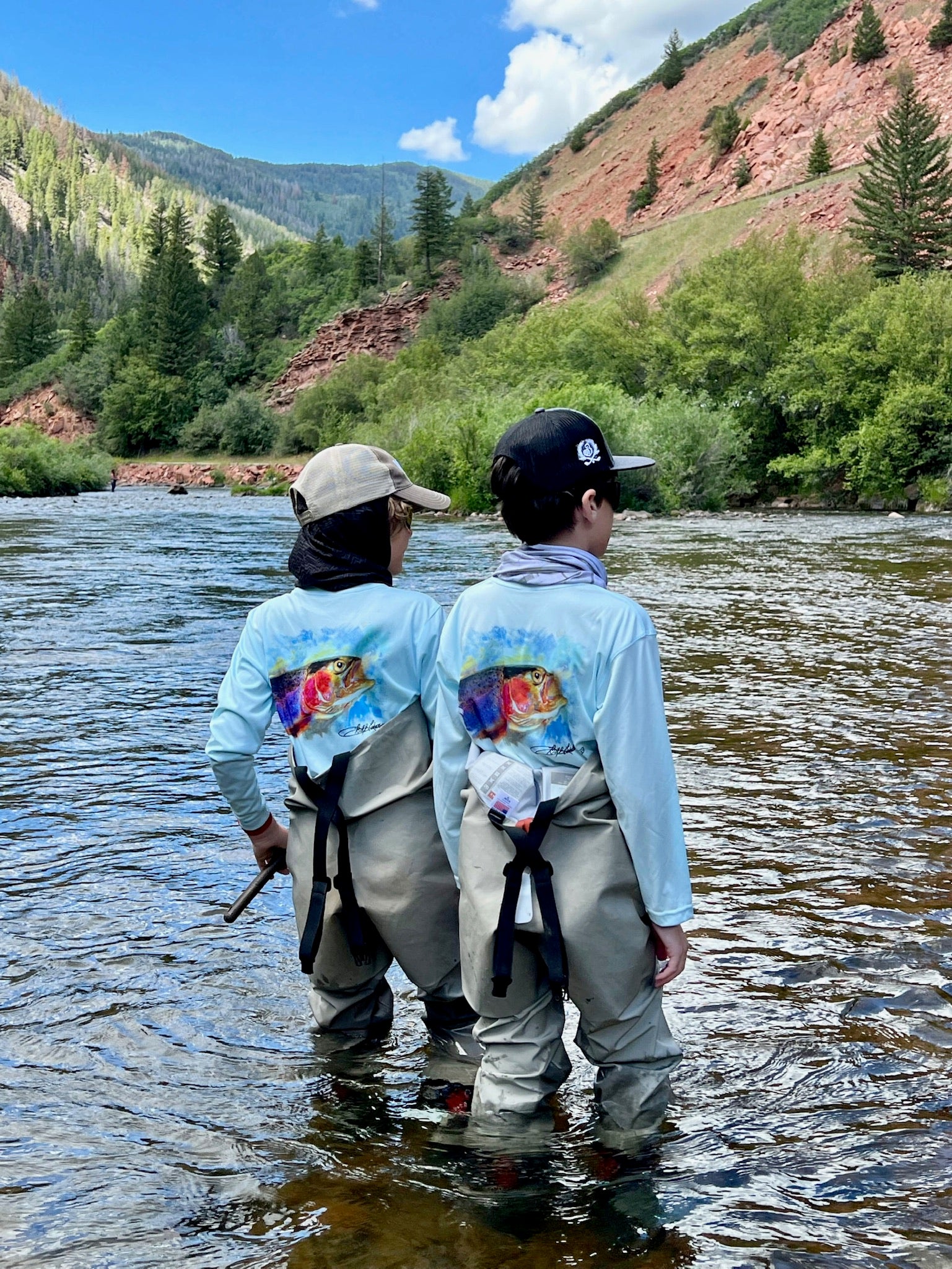 Two kids fly fishing in a river wearing Rocky Rainbow Trout Kids Performance Fishing Shirts with colorful trout print and long sleeve sun protection.