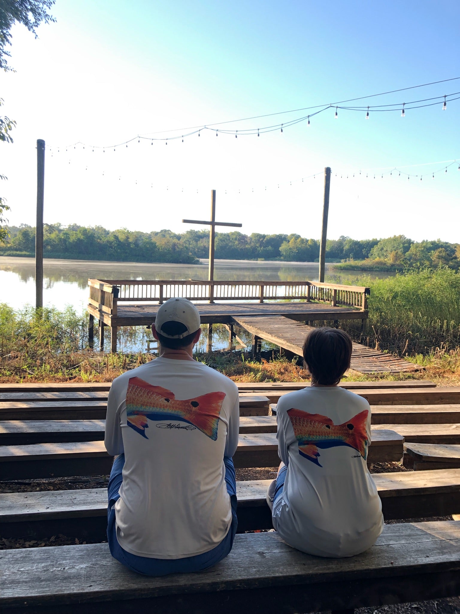 Two people wearing Redfish Design Fishing Shirt with UPF 50 sitting outdoors near a lake, showcasing the vibrant redfish graphic on the back.