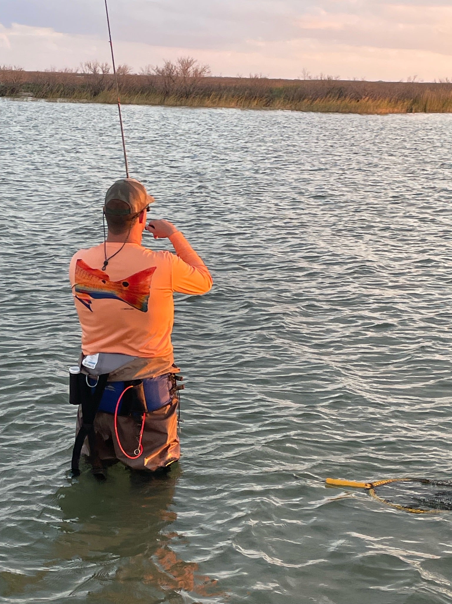 Angler wearing Orange Fishing Performance Shirt UPF 50+ with redfish tail design while wading in water, showcasing sun-protective fishing apparel in action.