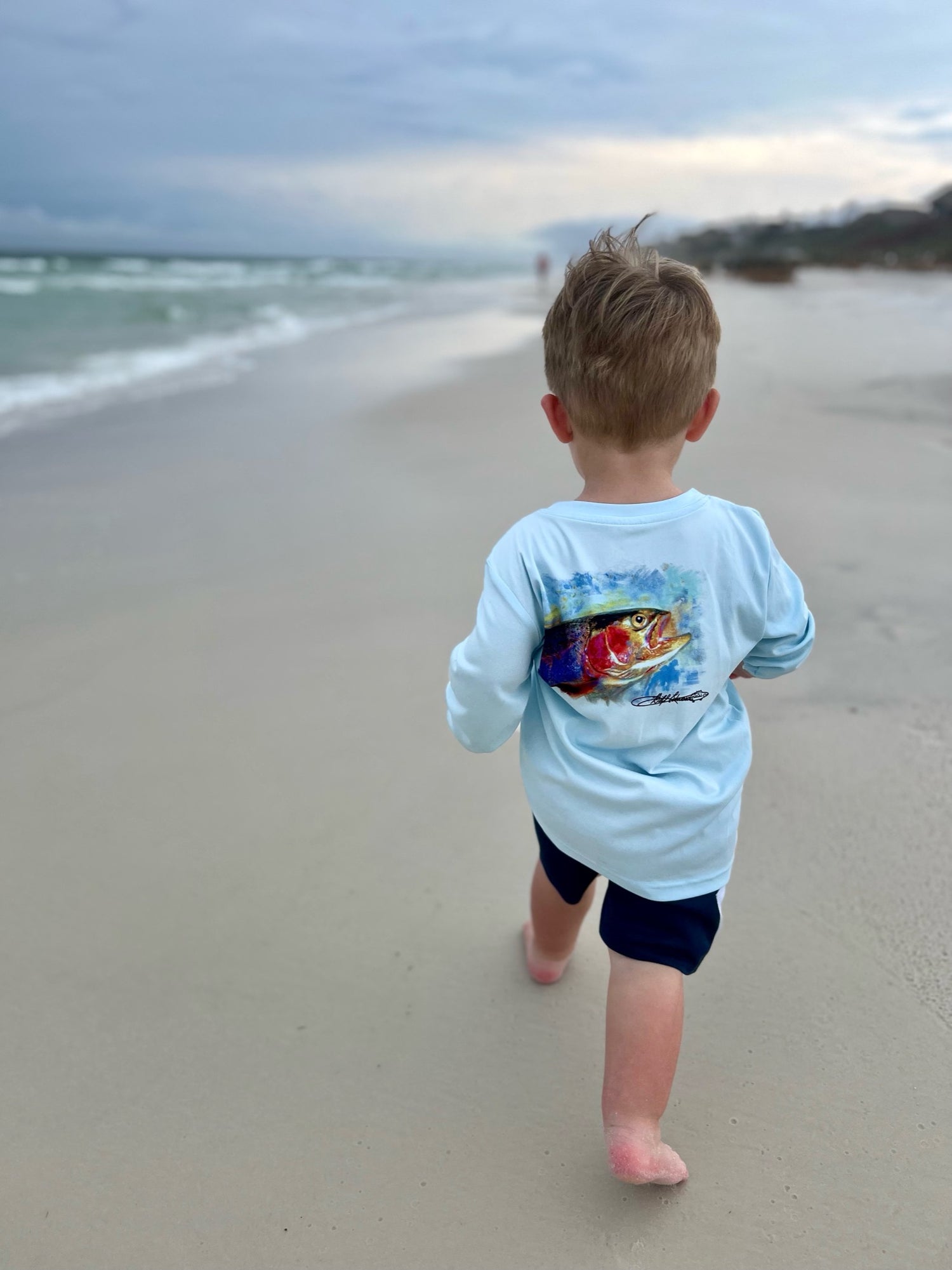 Young boy walking on beach in Rocky Rainbow Trout Kids Performance Fishing Shirt, designed for sun protection and moisture-wicking comfort.
