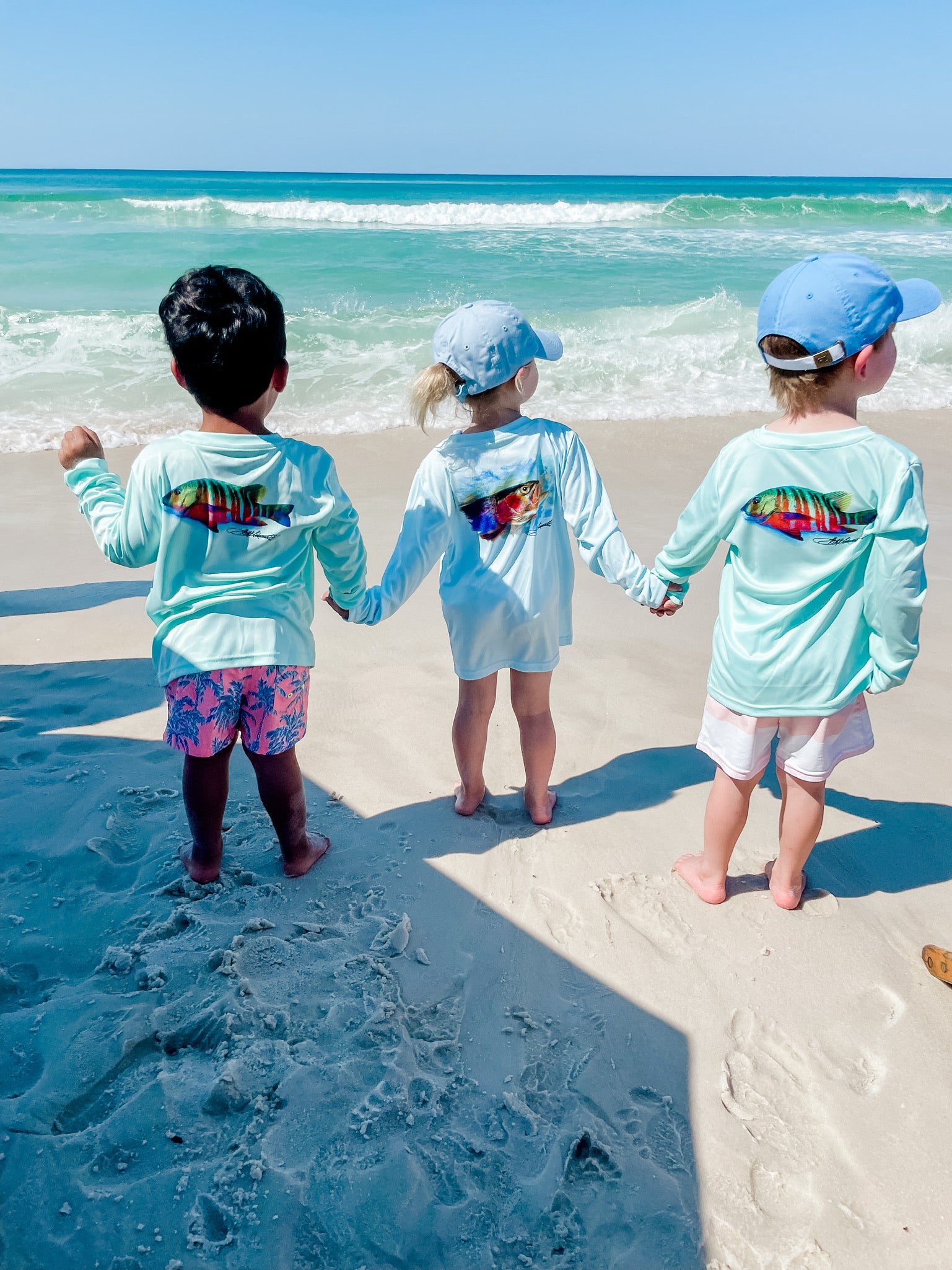 Kids holding hands on the beach wearing Santiago Snapper Performance Fishing Shirts UPF 50+, featuring colorful snapper art and sun-protective fabric.