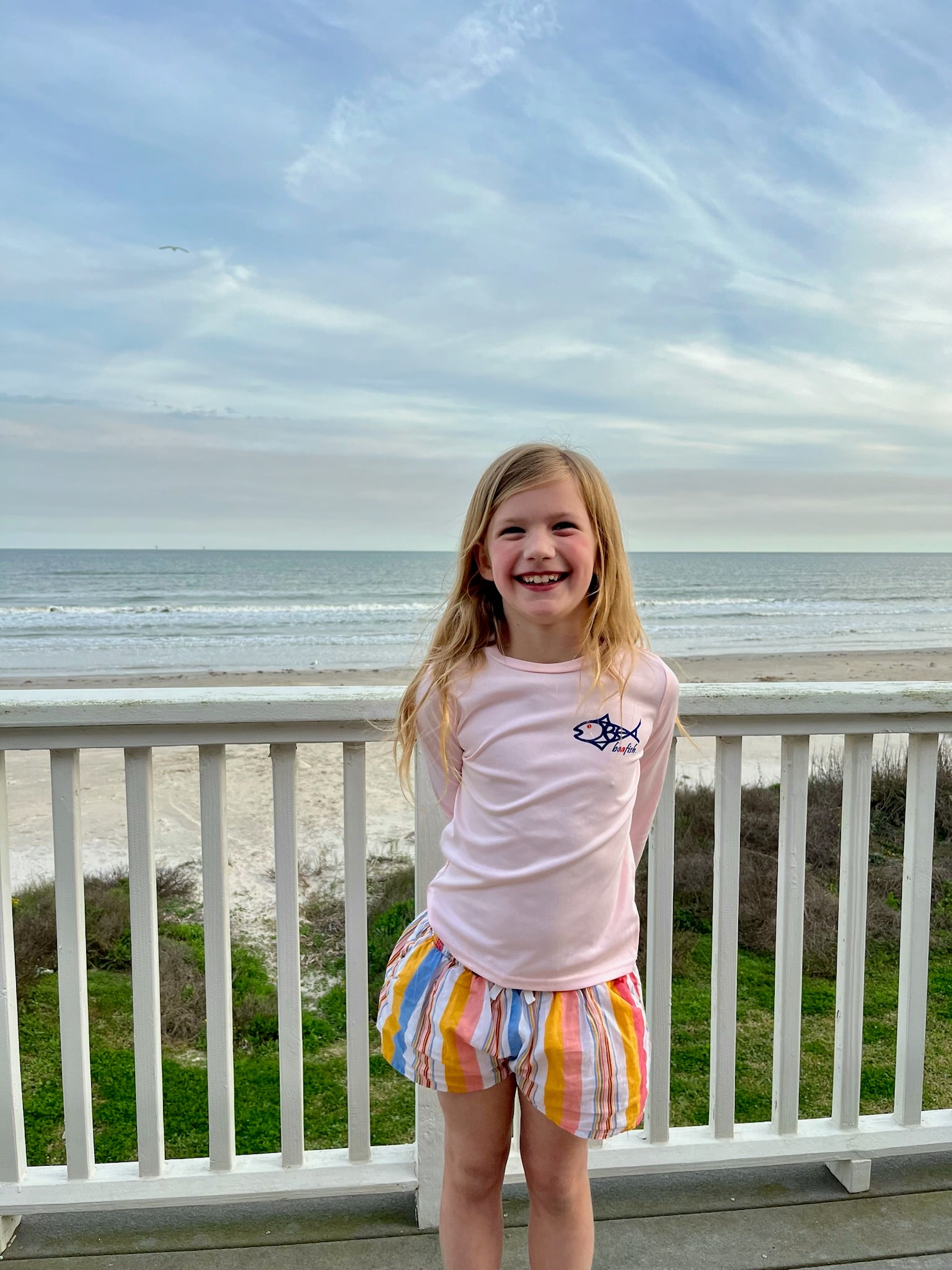 Smiling girl wearing Caribbean Iguana Kids Performance Fishing Shirt UPF 50+ with fish logo, standing on a beachside balcony.
