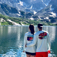 Couple wearing Rainbow Trout UPF 50+ Shirts by mountain lake, displaying matching trout artwork for outdoor couples' apparel.