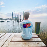 Man sitting on dock at sunset in Rainbow Trout UPF 50+ Shirt, highlighting back trout design and relaxed outdoor lifestyle appeal.
