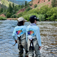Two kids fly fishing in a river wearing Rocky Rainbow Trout Kids Performance Fishing Shirts with colorful trout print and long sleeve sun protection.