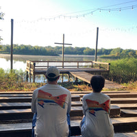 Two people wearing Redfish Design Fishing Shirt with UPF 50 sitting outdoors near a lake, showcasing the vibrant redfish graphic on the back.