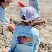 Toddler in sand wearing Rocky Rainbow Trout Kids Performance Fishing Shirt with vivid trout artwork, ideal for beach play and sun safety.