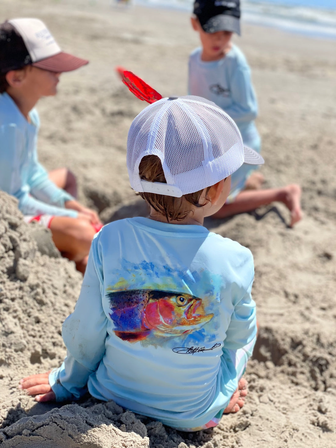 Toddler in sand wearing Rocky Rainbow Trout Kids Performance Fishing Shirt with vivid trout artwork, ideal for beach play and sun safety.