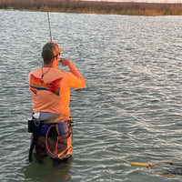 Angler wearing Orange Fishing Performance Shirt UPF 50+ with redfish tail design while wading in water, showcasing sun-protective fishing apparel in action.