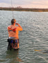 Angler wearing Orange Fishing Performance Shirt UPF 50+ with redfish tail design while wading in water, showcasing sun-protective fishing apparel in action.